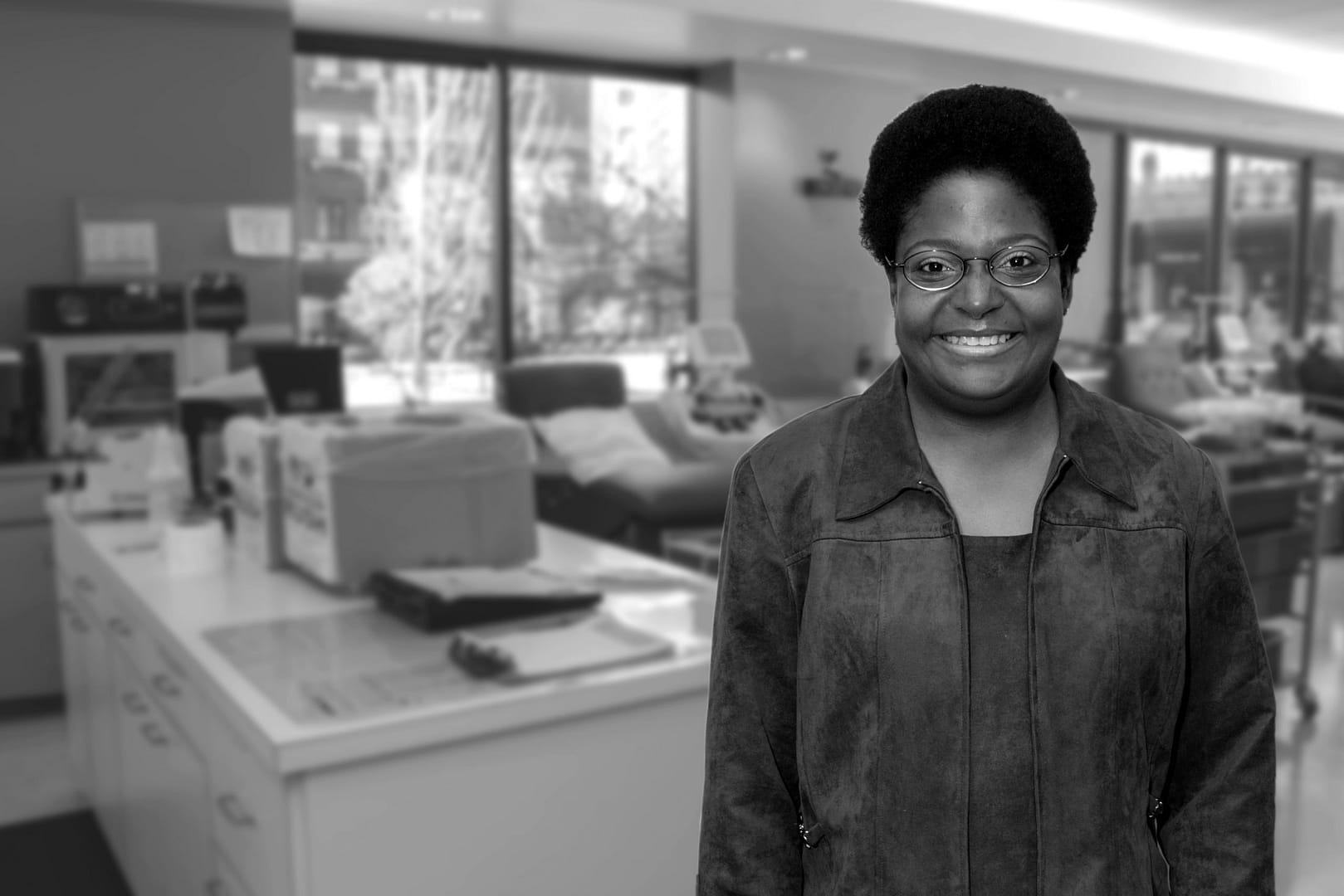 kendra Smiling Woman With Short Hair And Glasses Wearing A Dark Jacket, Standing In A Blood Donation Room With A Bed And Medical Equipment In The Background