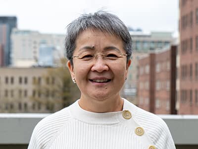 delight-resz Smiling Woman With Short Gray Hair Wearing Glasses And A White Sweater, Standing Outdoors With A Cityscape In The Background